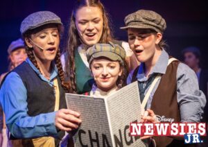 Four Newsies crowd around a newspaper with the word "change" written on the front.